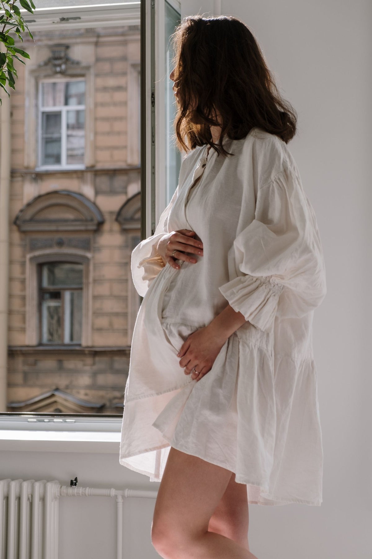 Pregnant woman in a white dress holding her belly beside a sunlit window