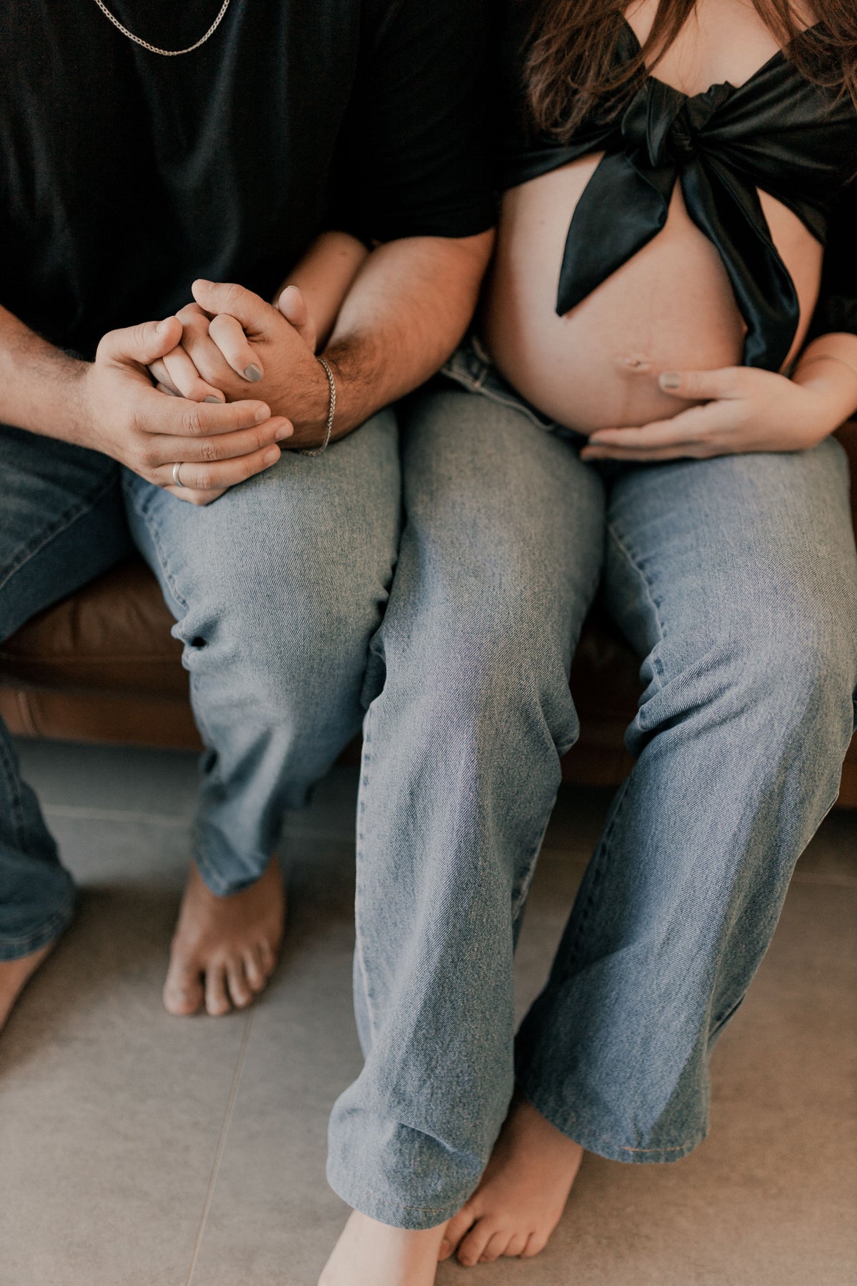 Pregnant woman and her partner sitting together on a sofa, holding hands beside her baby bump.